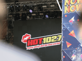 A stage display screen at Marks Park shows the HOT 102.7 FM logo with the text “Playing Joburg’s Best Old Skool and R&B.” The view is partially obscured by foreground objects and a colorful “NOW” banner to the right.