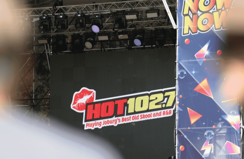 A stage display screen at Marks Park shows the HOT 102.7 FM logo with the text “Playing Joburg’s Best Old Skool and R&B.” The view is partially obscured by foreground objects and a colorful “NOW” banner to the right.