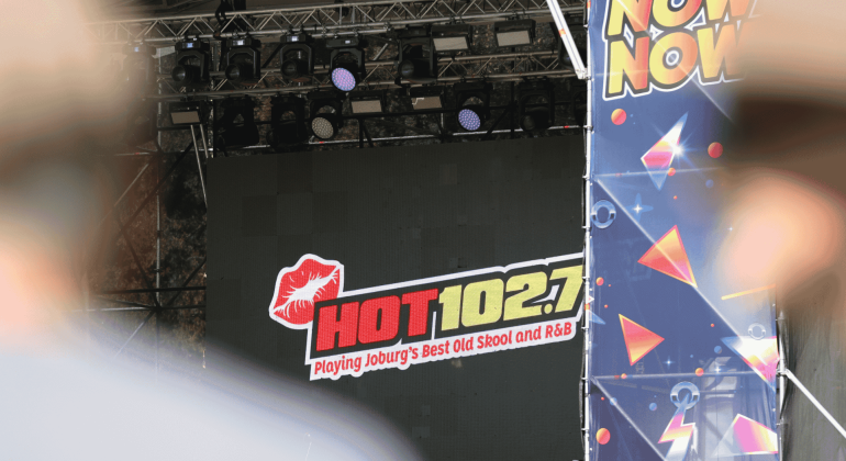 A stage display screen at Marks Park shows the HOT 102.7 FM logo with the text “Playing Joburg’s Best Old Skool and R&B.” The view is partially obscured by foreground objects and a colorful “NOW” banner to the right.