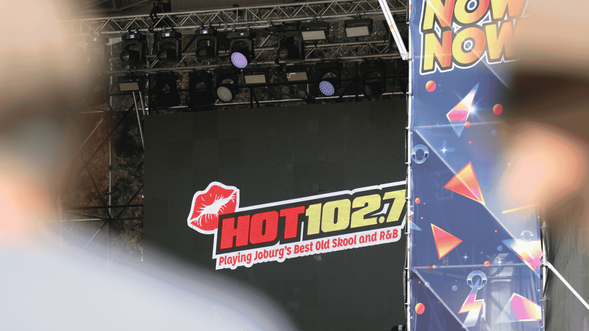 A stage display screen at Marks Park shows the HOT 102.7 FM logo with the text “Playing Joburg’s Best Old Skool and R&B.” The view is partially obscured by foreground objects and a colorful “NOW” banner to the right.