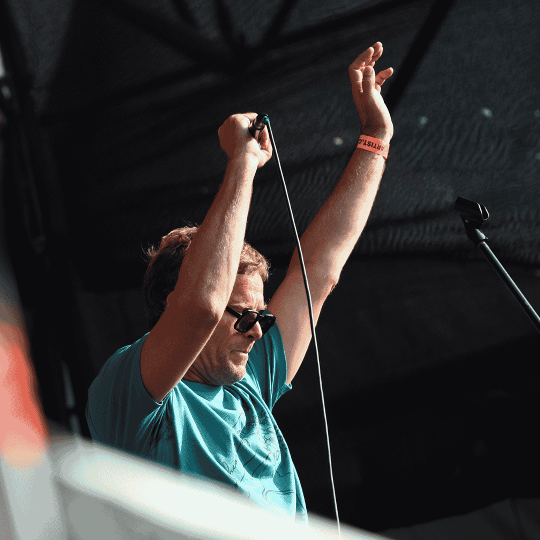 A man wearing sunglasses and a blue t-shirt raises both arms while holding a microphone on stage, performing under an outdoor canopy at Marks Park.