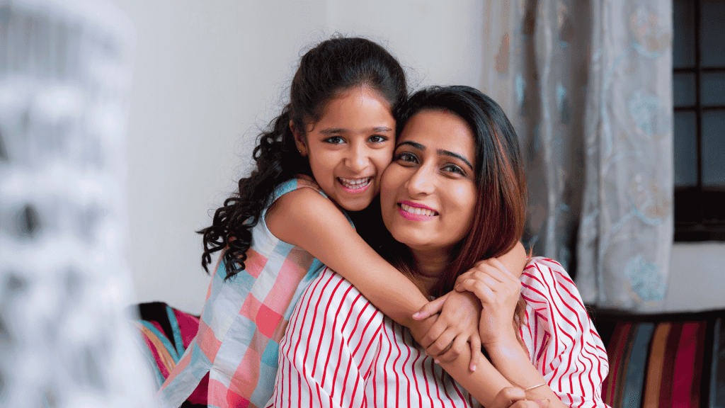 A smiling young girl with curly hair hugs a woman, likely her mother, from behind. Both are indoors on a cozy couch, radiating happiness and the true wealth of family bonds. Soft curtains and part of a window appear in the background.
