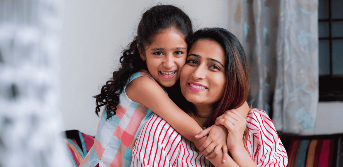 A smiling young girl with curly hair hugs a woman, likely her mother, from behind. Both are indoors on a cozy couch, radiating happiness and the true wealth of family bonds. Soft curtains and part of a window appear in the background.