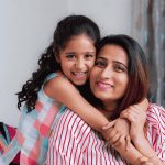 A smiling young girl with curly hair hugs a woman, likely her mother, from behind. Both are indoors on a cozy couch, radiating happiness and the true wealth of family bonds. Soft curtains and part of a window appear in the background.