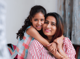A smiling young girl with curly hair hugs a woman, likely her mother, from behind. Both are indoors on a cozy couch, radiating happiness and the true wealth of family bonds. Soft curtains and part of a window appear in the background.