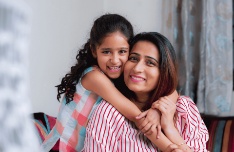 A smiling young girl with curly hair hugs a woman, likely her mother, from behind. Both are indoors on a cozy couch, radiating happiness and the true wealth of family bonds. Soft curtains and part of a window appear in the background.