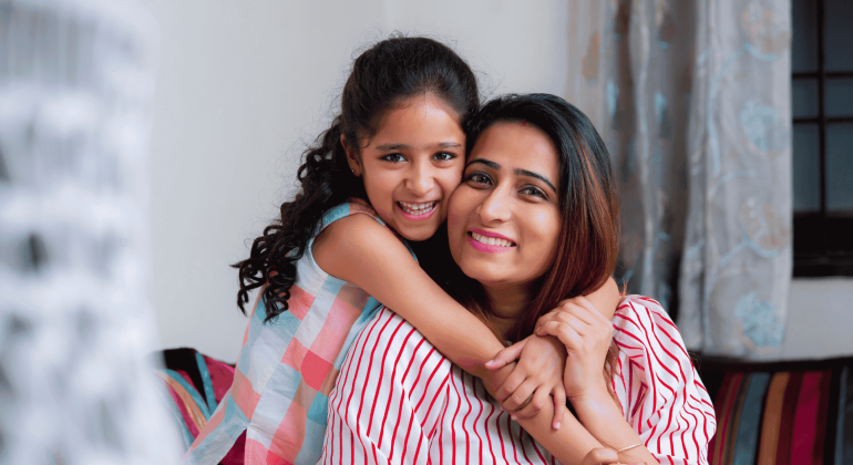 A smiling young girl with curly hair hugs a woman, likely her mother, from behind. Both are indoors on a cozy couch, radiating happiness and the true wealth of family bonds. Soft curtains and part of a window appear in the background.