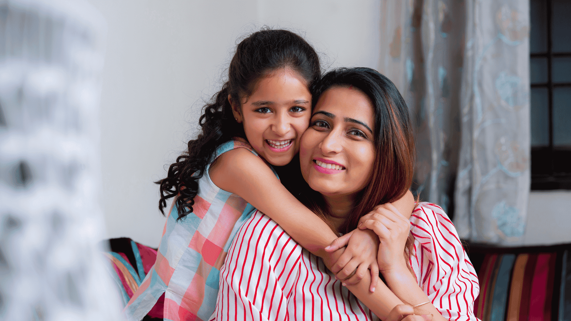 A smiling young girl with curly hair hugs a woman, likely her mother, from behind. Both are indoors on a cozy couch, radiating happiness and the true wealth of family bonds. Soft curtains and part of a window appear in the background.