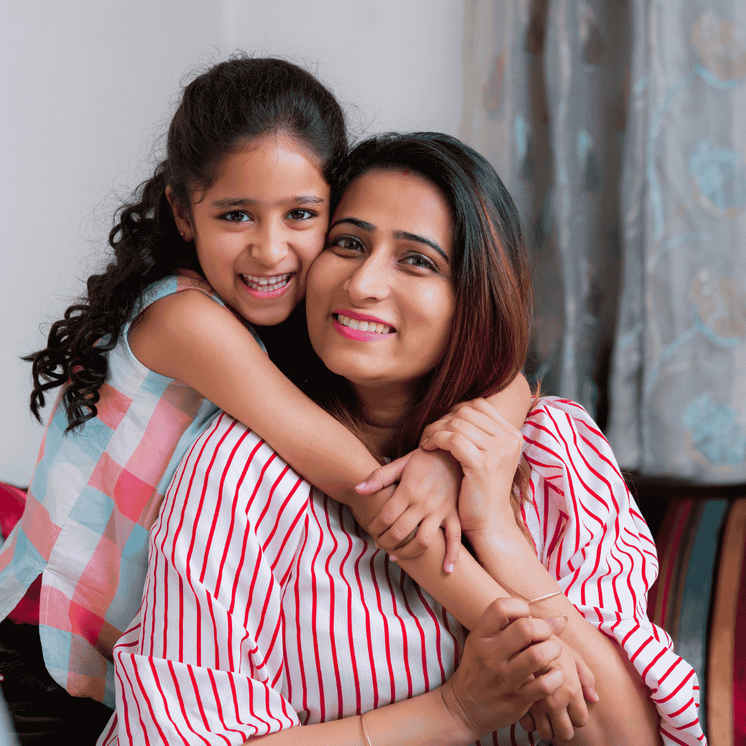 A smiling woman sits while a young girl hugs her from behind. Both look happy, wearing striped tops that hint at simple wealth. The background shows curtains and a soft, cozy indoor setting.
