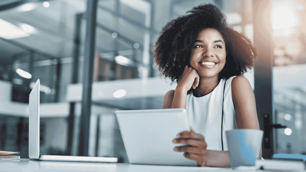 A woman sits at a desk in a modern office, smiling while holding a tablet. A laptop and coffee mug are on the desk. She looks thoughtfully out of the frame, sunlight spilling in and reflecting an air of wealth and success.