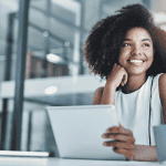 A woman sits at a desk in a modern office, smiling while holding a tablet. A laptop and coffee mug are on the desk. She looks thoughtfully out of the frame, sunlight spilling in and reflecting an air of wealth and success.