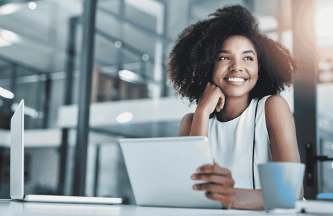 A woman sits at a desk in a modern office, smiling while holding a tablet. A laptop and coffee mug are on the desk. She looks thoughtfully out of the frame, sunlight spilling in and reflecting an air of wealth and success.