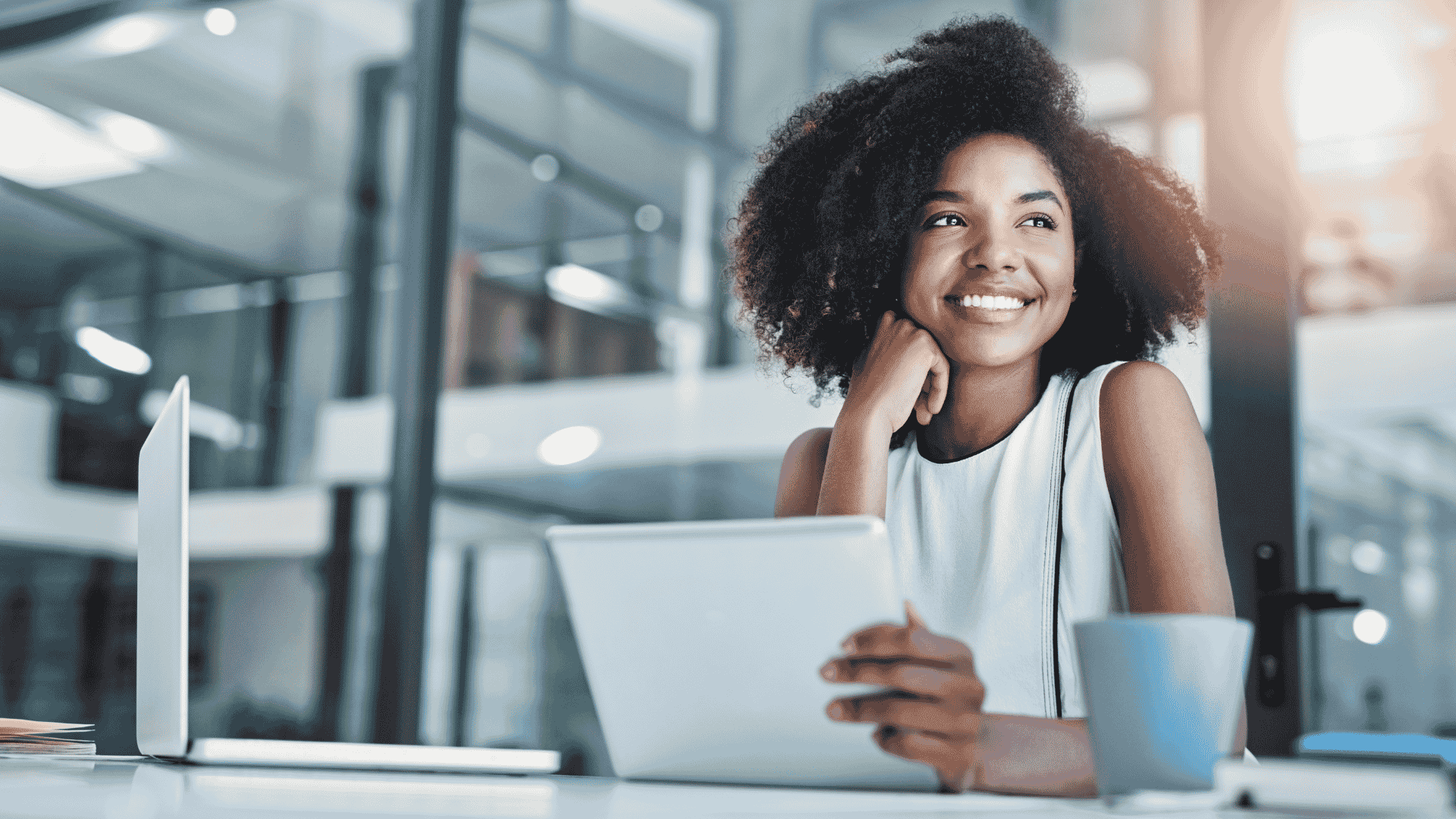 A woman sits at a desk in a modern office, smiling while holding a tablet. A laptop and coffee mug are on the desk. She looks thoughtfully out of the frame, sunlight spilling in and reflecting an air of wealth and success.