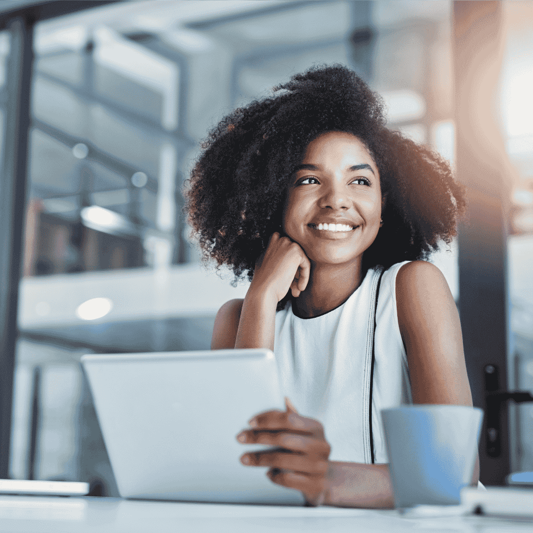 A woman with curly hair sits at a desk, smiling and holding a tablet. She is wearing a sleeveless top, with a coffee cup in front of her, exuding confidence and wealth in a modern, bright office space with glass walls.