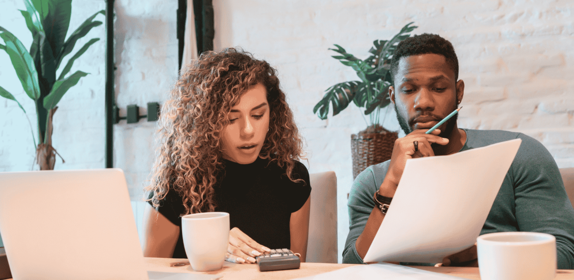 A woman and a man sit at a desk with a laptop, coffee mugs, papers, and a calculator, appearing focused as they review documents related to wealth together in a bright, modern office space with plants in the background.