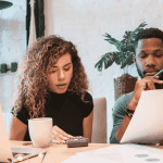 A woman and a man sit at a desk with a laptop, coffee mugs, papers, and a calculator, appearing focused as they review documents related to wealth together in a bright, modern office space with plants in the background.