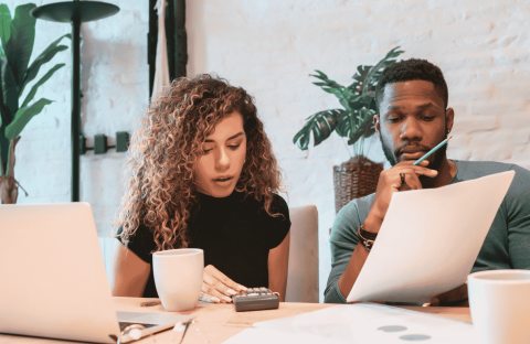 A woman and a man sit at a desk with a laptop, coffee mugs, papers, and a calculator, appearing focused as they review documents related to wealth together in a bright, modern office space with plants in the background.