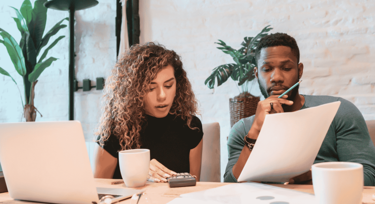 A woman and a man sit at a desk with a laptop, coffee mugs, papers, and a calculator, appearing focused as they review documents related to wealth together in a bright, modern office space with plants in the background.
