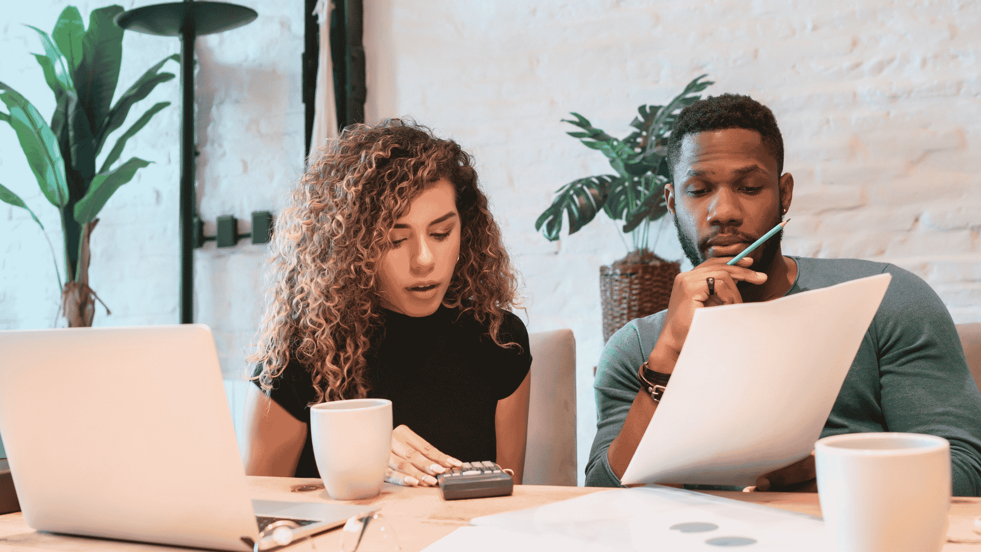 A woman and a man sit at a desk with a laptop, coffee mugs, papers, and a calculator, appearing focused as they review documents related to wealth together in a bright, modern office space with plants in the background.