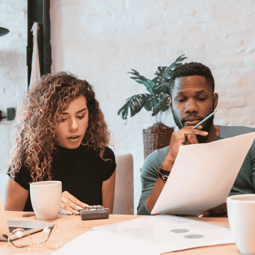 Two people sit at a table with coffee cups, working together. Focused and engaged, they discuss their finances—one uses a calculator while the other reviews a sheet of paper, both intent on managing their wealth.