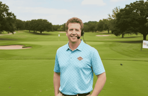 A man in a blue polo shirt and headset stands smiling on a green golf course, with trees, sand traps, and a golf flag visible in the background. A Hot 1027 Breakfast sign is seen on the right side of the image.