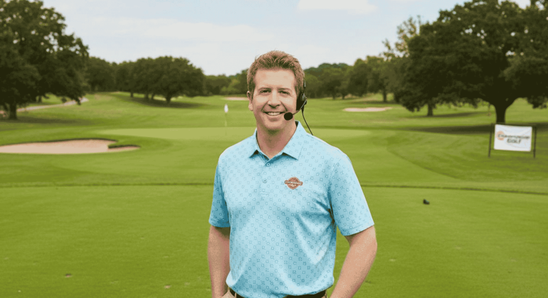 A man in a blue polo shirt and headset stands smiling on a green golf course, with trees, sand traps, and a golf flag visible in the background. A Hot 1027 Breakfast sign is seen on the right side of the image.