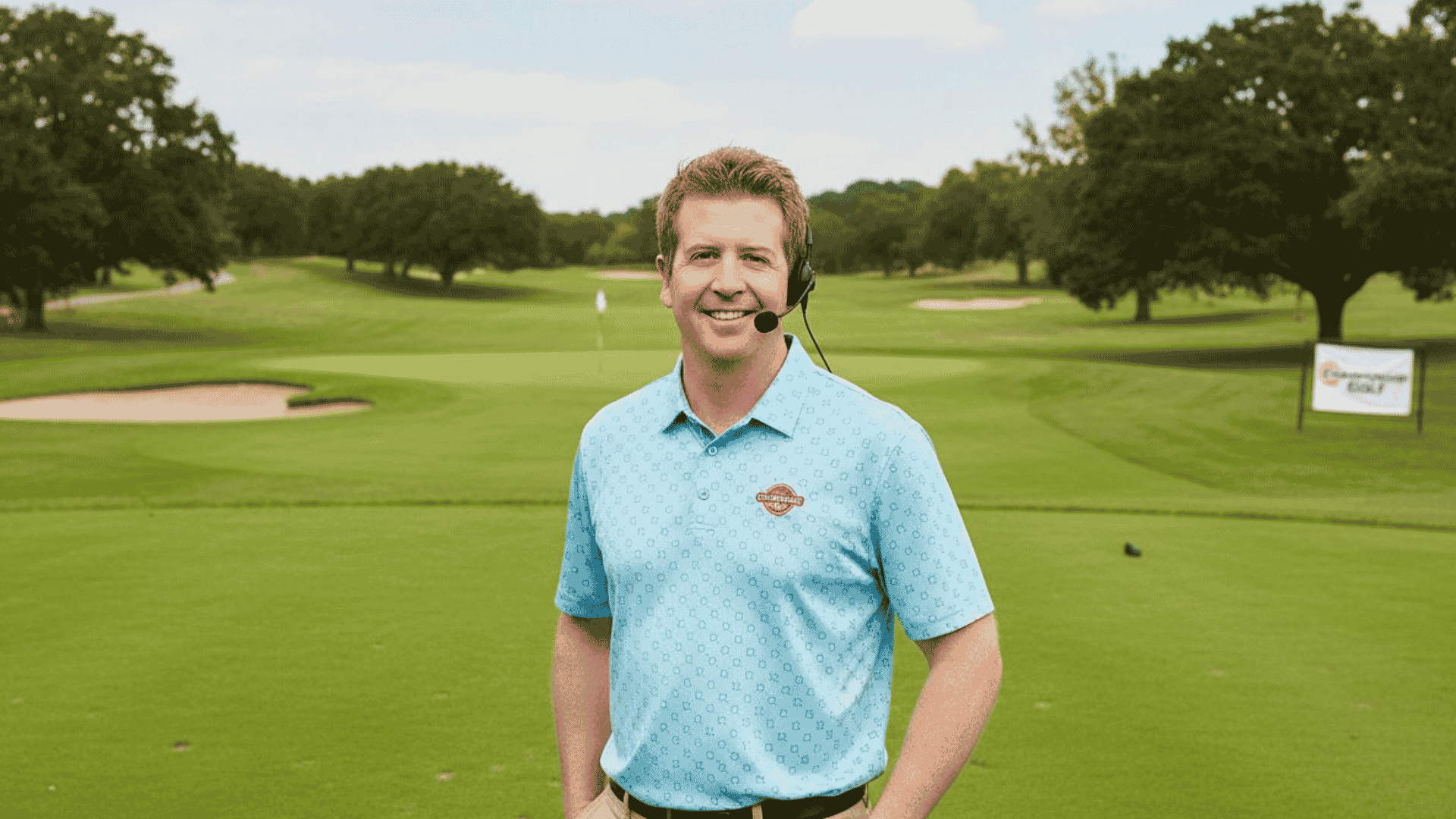 A man in a blue polo shirt and headset stands smiling on a green golf course, with trees, sand traps, and a golf flag visible in the background. A Hot 1027 Breakfast sign is seen on the right side of the image.