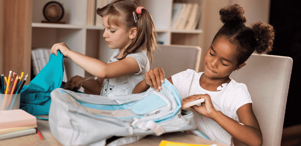 Two young girls sit at a table, focused on organizing their backpacks. One has light hair in pigtails, the other dark curly hair in buns. School supplies, books, and colored pencils fill the space—like a scene from an Ali Campbell story.