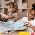Two young girls sit at a table, focused on organizing their backpacks. One has light hair in pigtails, the other dark curly hair in buns. School supplies, books, and colored pencils fill the space—like a scene from an Ali Campbell story.