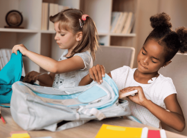 Two young girls sit at a table, focused on organizing their backpacks. One has light hair in pigtails, the other dark curly hair in buns. School supplies, books, and colored pencils fill the space—like a scene from an Ali Campbell story.