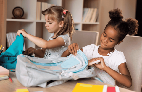 Two young girls sit at a table, focused on organizing their backpacks. One has light hair in pigtails, the other dark curly hair in buns. School supplies, books, and colored pencils fill the space—like a scene from an Ali Campbell story.