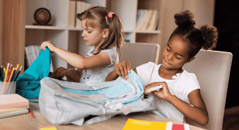 Two young girls sit at a table, focused on organizing their backpacks. One has light hair in pigtails, the other dark curly hair in buns. School supplies, books, and colored pencils fill the space—like a scene from an Ali Campbell story.