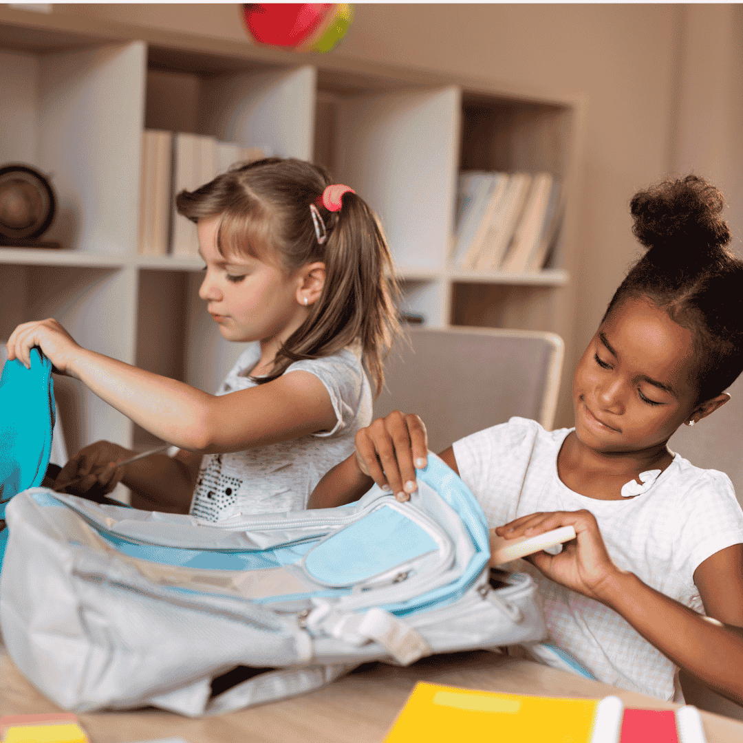 Two young girls are sitting at a table, packing their backpacks with books and supplies. They appear focused, as if preparing for a big day—maybe even listening to Ali Campbell while surrounded by shelves filled with books in the background.