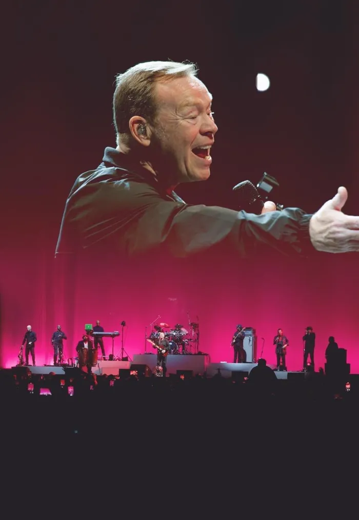 A band performs on stage with pink lighting, while a large screen above shows a close-up of the lead singer passionately singing. Audience silhouettes, eager to soak in the wealth of talent, are visible in the foreground.