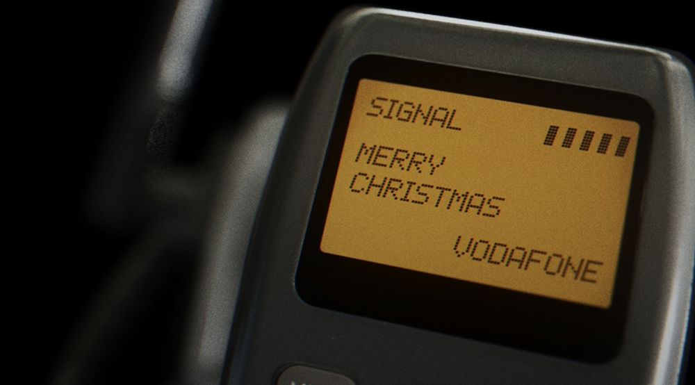 A close-up of an old mobile phone screen displaying the words MERRY CHRISTMAS above the Vodafone network name and a full signal bar indicator, reminiscent of texting friends about top hits, with a dark, blurred background.