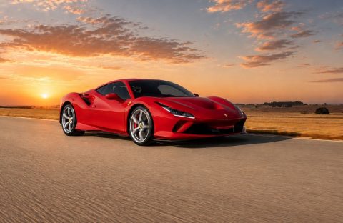 A red Ferrari Club sports car drives on an open road at sunset, with dramatic clouds and a golden sky in the background. The flat, grassy landscape highlights the sleek car&rsquo;s vibrant color and dynamic presence.