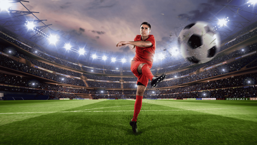Lloyd Madurai, in a red uniform, kicks a ball mid-air in a brightly lit stadium filled with spectators, with the large ball appearing close to the camera.