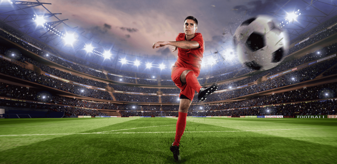 Lloyd Madurai, in a red uniform, kicks a ball mid-air in a brightly lit stadium filled with spectators, with the large ball appearing close to the camera.