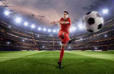 Lloyd Madurai, in a red uniform, kicks a ball mid-air in a brightly lit stadium filled with spectators, with the large ball appearing close to the camera.