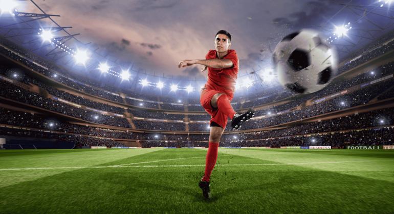 Lloyd Madurai, in a red uniform, kicks a ball mid-air in a brightly lit stadium filled with spectators, with the large ball appearing close to the camera.