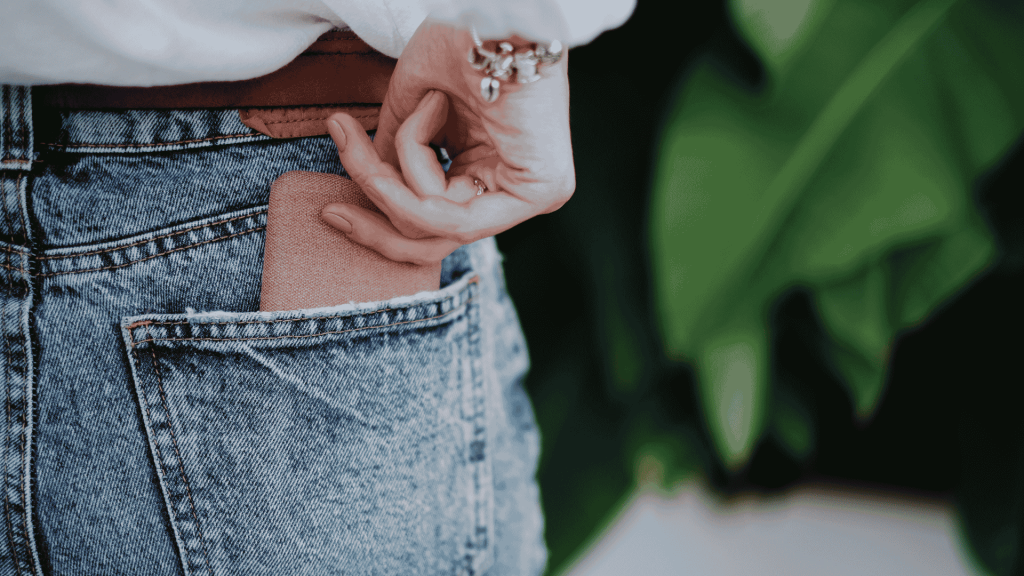 A person wearing blue denim jeans is placing a brown wallet into their back pocket. Their hand is partially visible, and they are wearing a white top and a bracelet. Green leaves are blurred in the background.