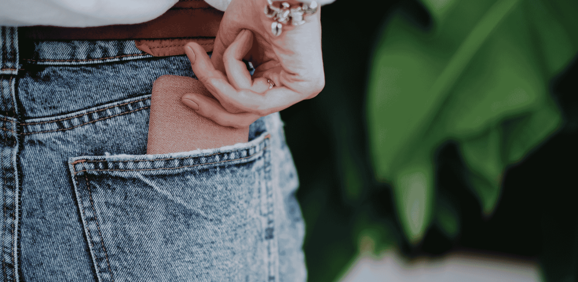 A person wearing blue denim jeans is placing a brown wallet into their back pocket. Their hand is partially visible, and they are wearing a white top and a bracelet. Green leaves are blurred in the background.