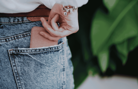 A person wearing blue denim jeans is placing a brown wallet into their back pocket. Their hand is partially visible, and they are wearing a white top and a bracelet. Green leaves are blurred in the background.