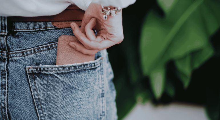 A person wearing blue denim jeans is placing a brown wallet into their back pocket. Their hand is partially visible, and they are wearing a white top and a bracelet. Green leaves are blurred in the background.