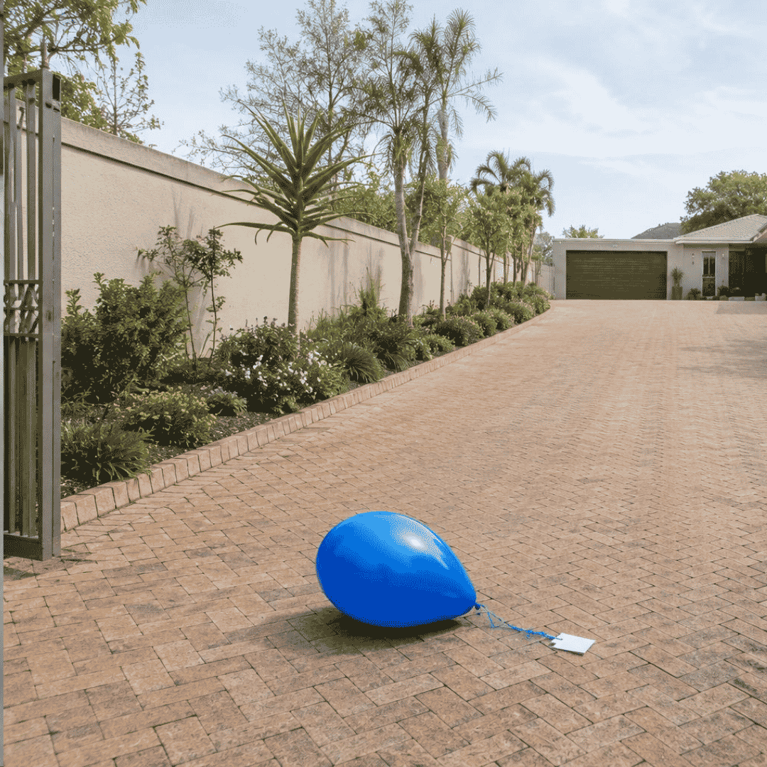 A blue balloon with a string and a small white card attached lies on a brick driveway beside a garden with palm trees, where the breeze feels like it could have drifted in from two oceans, leading up to a closed garage and a house in the background.