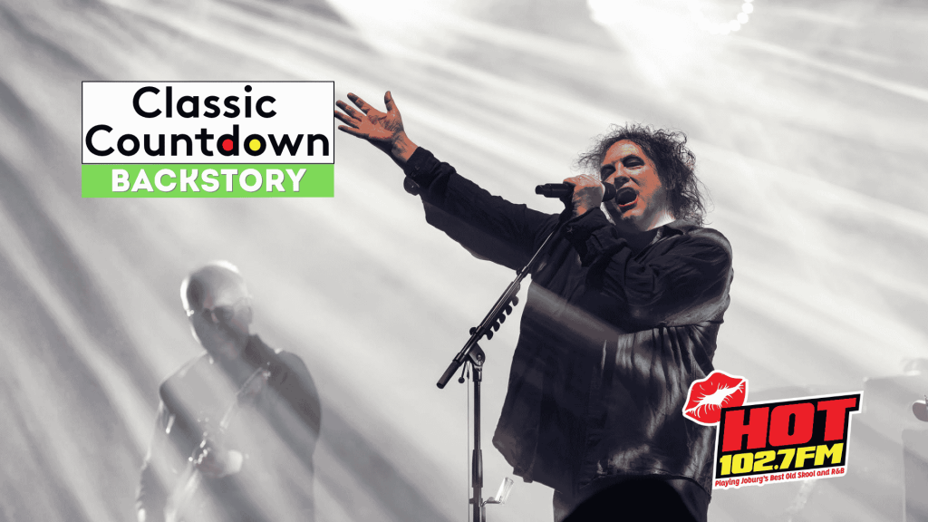 A singer with curly hair performs passionately on stage under dramatic lighting, with a guitarist in the background. The image features the Classic Countdown Backstory and HOT 102.7 FM logos, evoking the energy of a Robert Palmer concert.