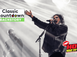 A singer with curly hair performs passionately on stage under dramatic lighting, with a guitarist in the background. The image features the Classic Countdown Backstory and HOT 102.7 FM logos, evoking the energy of a Robert Palmer concert.