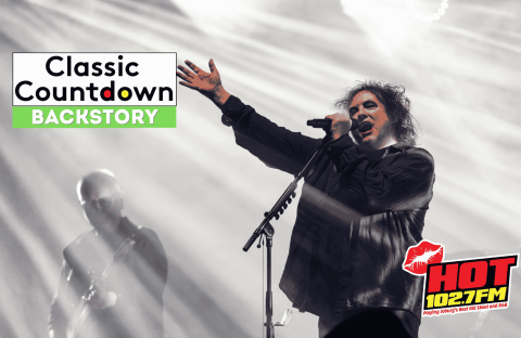 A singer with curly hair performs passionately on stage under dramatic lighting, with a guitarist in the background. The image features the Classic Countdown Backstory and HOT 102.7 FM logos, evoking the energy of a Robert Palmer concert.