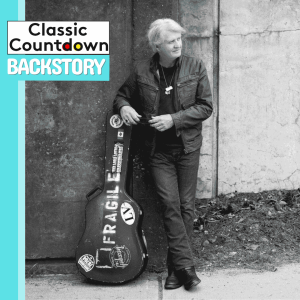 A man with light hair in dark clothing stands outdoors, leaning on a worn guitar case labeled FRAGILE. The text reads Classic Countdown Backstory in the top left corner, hinting at Robert Palmer&rsquo;s musical journey, on a blue and white background.