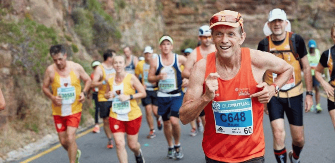 A group of marathon runners, wearing race bibs and colorful athletic gear, run on a winding road beside a rocky hillside. The man in front, smiling, wears an orange tank top and blue bib labeled C648.